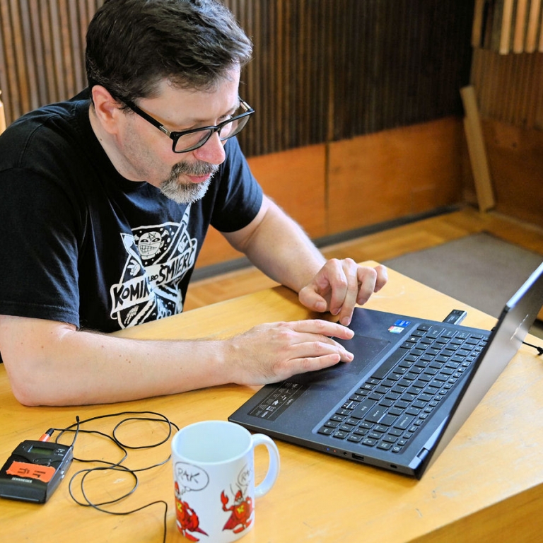 Jarosław Ejsymont siedzi przy stole przed laptopem, obok sprzęt nagrywający leży na stole