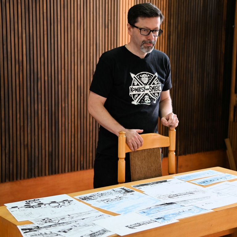Jarosław Ejsymont w czarnej koszulce z napisem Komiksy albo śmierć stoi przed stołem z rozłożonymi planszami komiksowymi