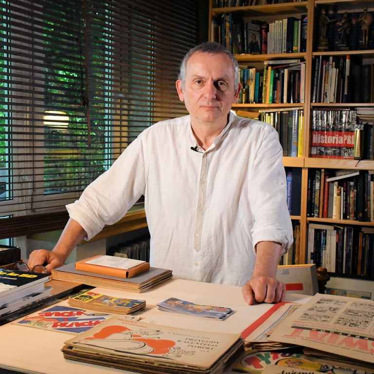 Wojciech Birek oparty o stół wraz ze zbiorami komiksowymi