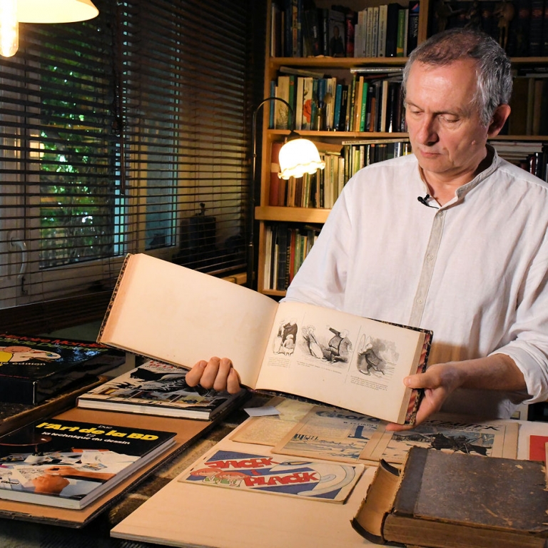 Wojciech Jama prezentuje scenkę komiksową w jednym w komiksów przed nim na stole leży dużo rozłożonych komiksów